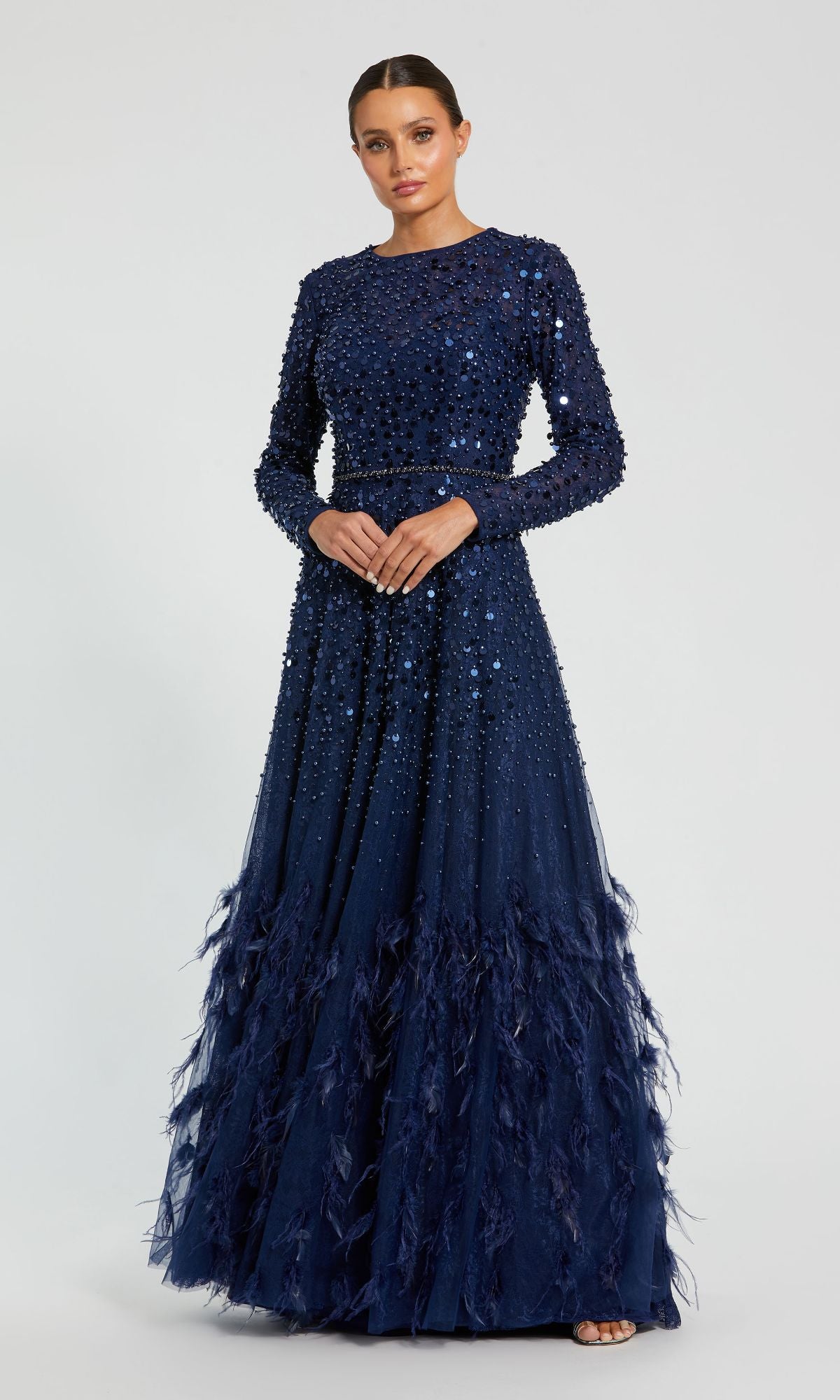 A woman wears the Mac Duggal 11782 Long Sleeve Sequin Formal Gown in navy blue, featuring beaded and sequin embellishments with feathered hem details—an elegant mother of the bride dress against a simple background.